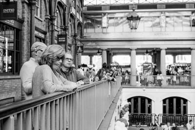 People lean on a balcony railing inside a busy London market hall, looking down at the lower level.
