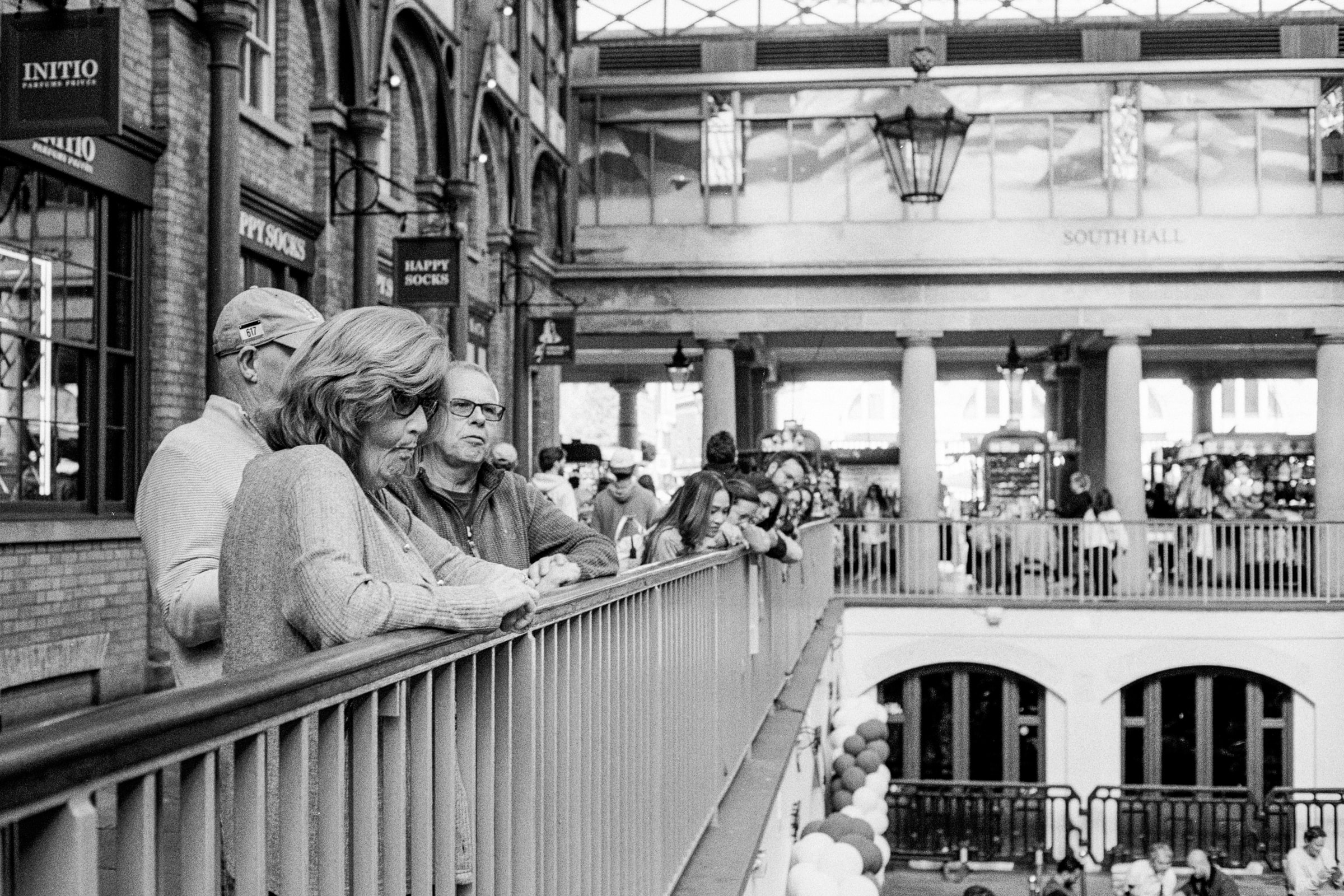 People lean on a balcony railing inside a busy London market hall, looking down at the lower level.