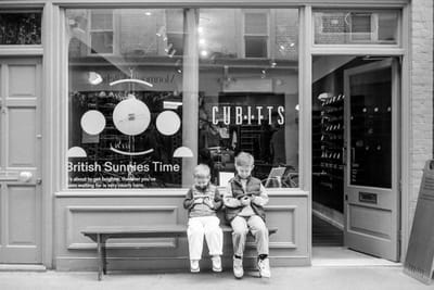 Two children sit on a bench outside a London shop window, both looking down at handheld devices.
