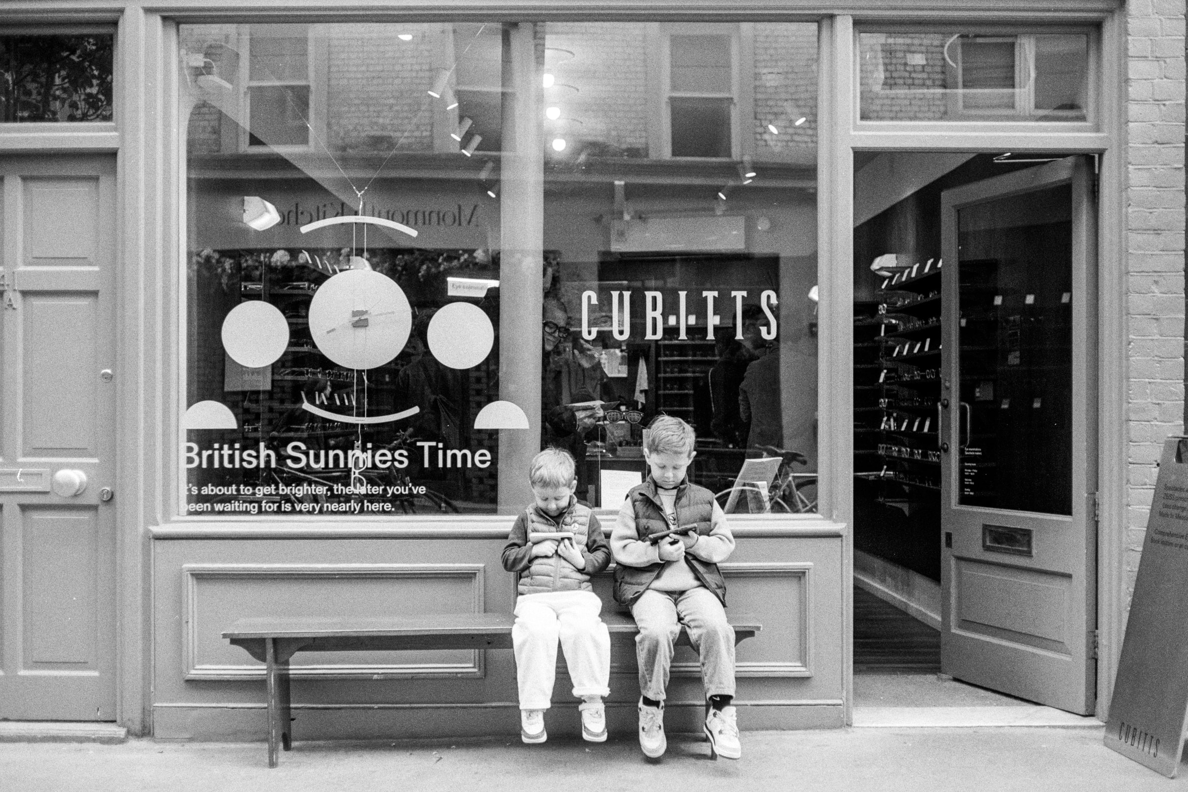 Two children sit on a bench outside a London shop window, both looking down at handheld devices.