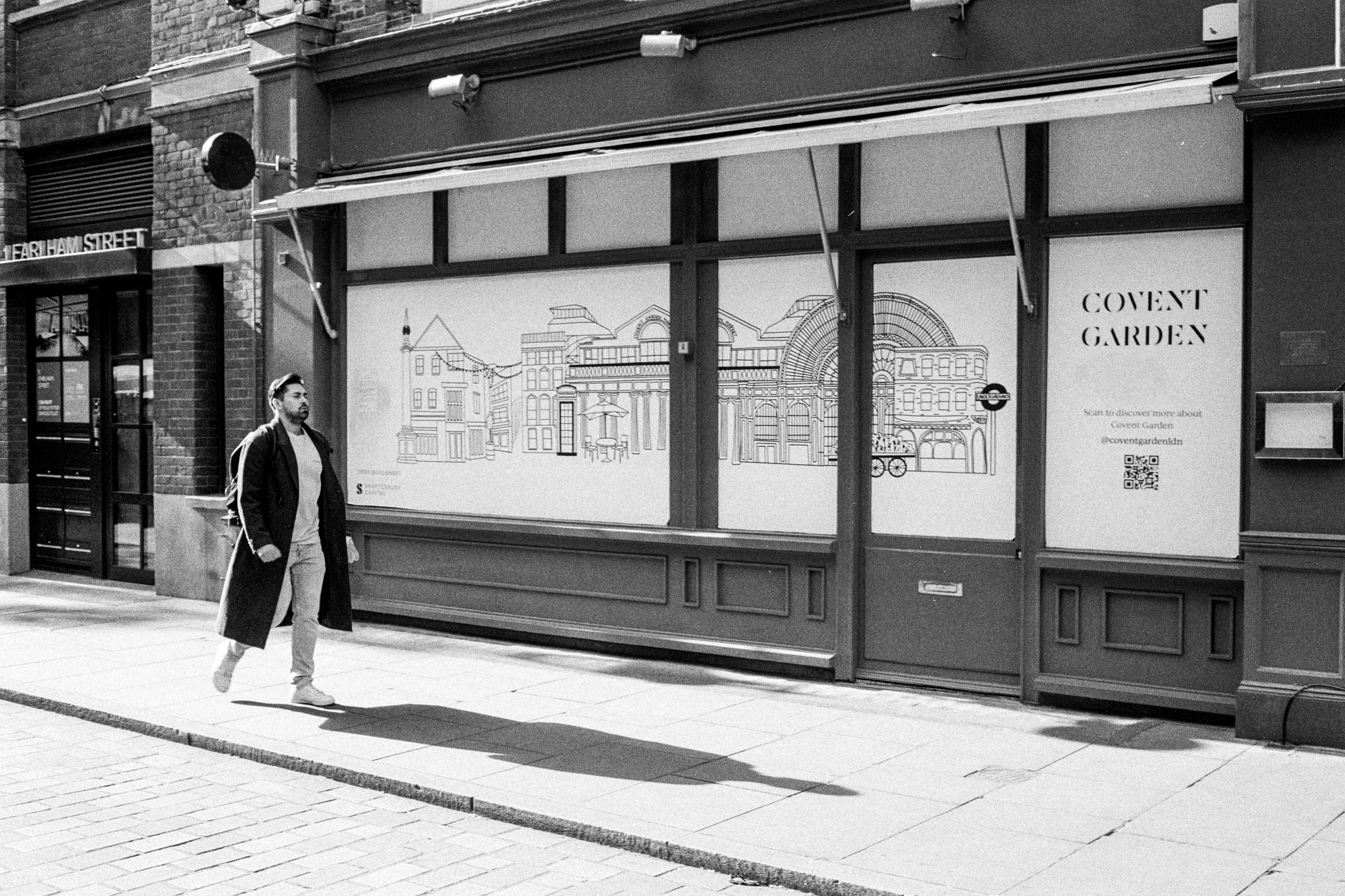 Man in a long coat walking past a Covent Garden storefront with illustrated window coverings in London.