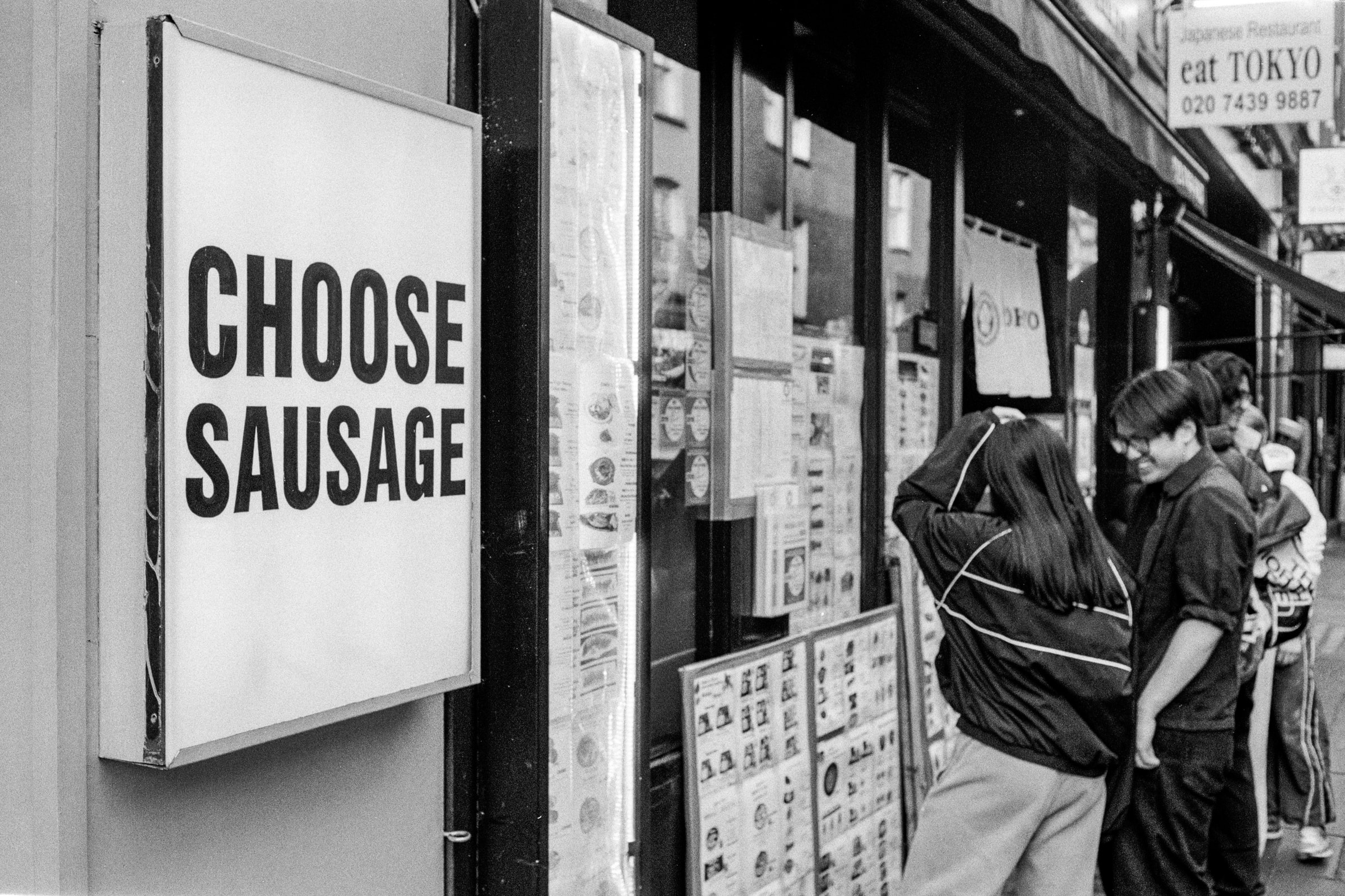 People stand outside a London restaurant beside a sign reading “CHOOSE SAUSAGE”