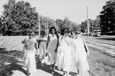 Six women in light summer dresses walk along a sunny sidewalk beside tram tracks in Riga.