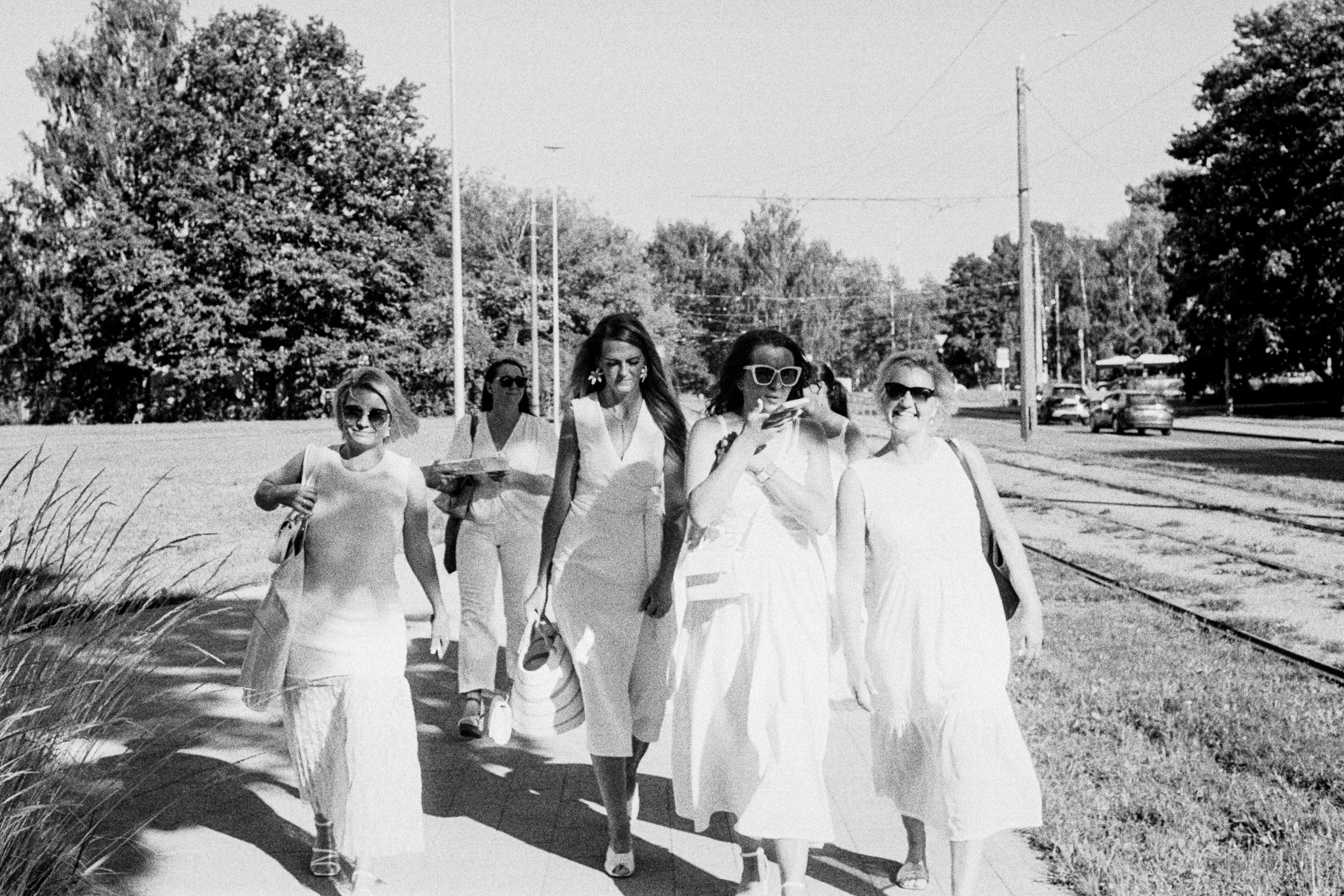 Six women in light summer dresses walk along a sunny sidewalk beside tram tracks in Riga.