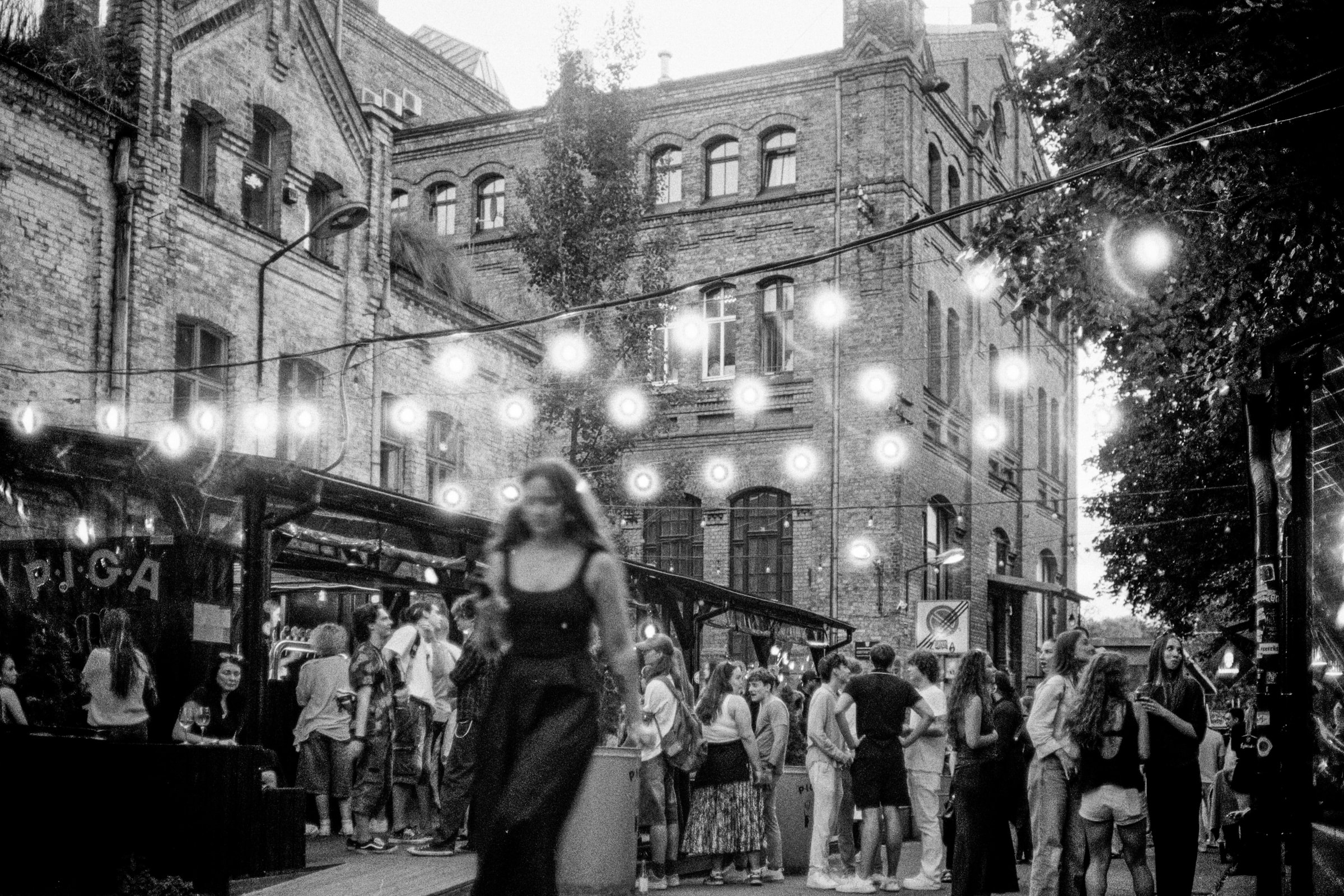 A crowd under string lights in a brick courtyard in Riga as a woman walks past in the foreground.